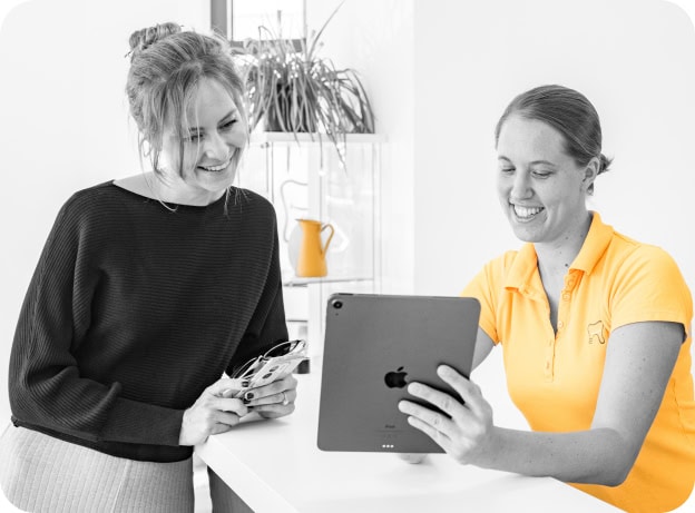 Zwei Frauen lächeln und betrachten ein Tablet in heller, moderner Einrichtung. Eine trägt ein gelbes Shirt.