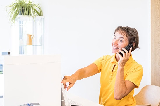 Mitarbeiterin am Schreibtisch telefoniert freudig, zeigt auf Computerbildschirm im Büro.